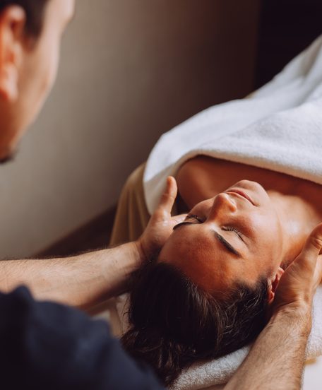 Eine Frau liegt auf einer Massageliege und erhält eine Kopf- und Nackenmassage in einem Wellnessraum.