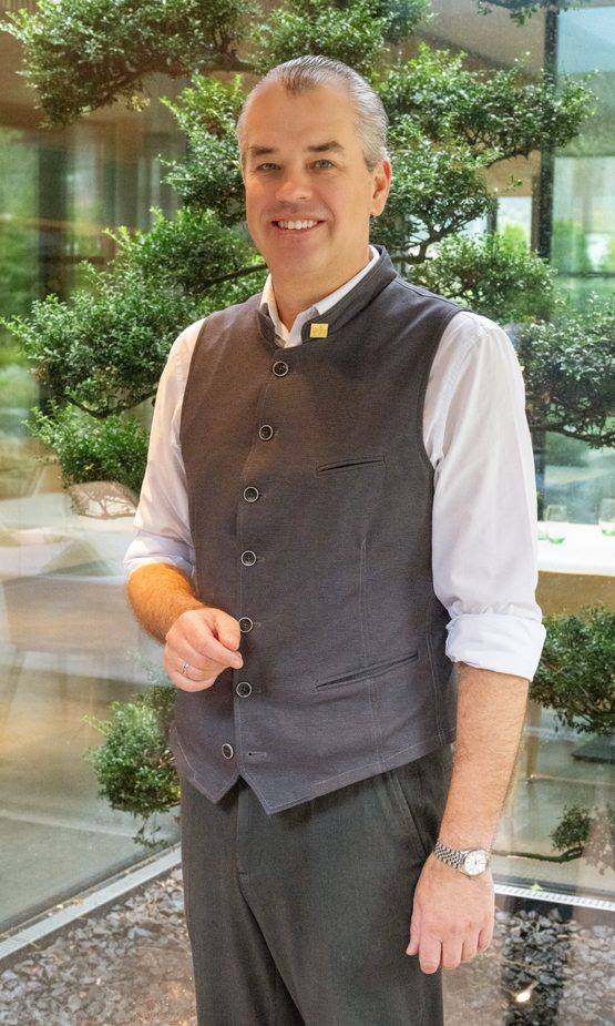 Ein Hotelmitarbeiter steht lächelnd im Innenbereich vor einer Glasfront und dekorativen Pflanzen.
