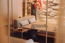 Wellness-Ruheraum mit gepolsterten Liegen, Natursteinwand, Beistelltisch und rosa Blüten in einer Vase.