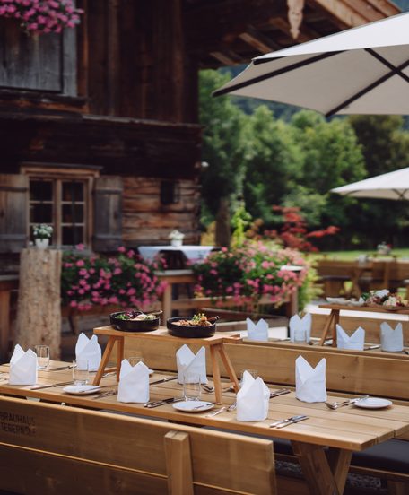 Gedeckte Holztische auf einer Restaurantterrasse vor einem rustikalen Holzhaus mit Blumen und Sonnenschirmen.