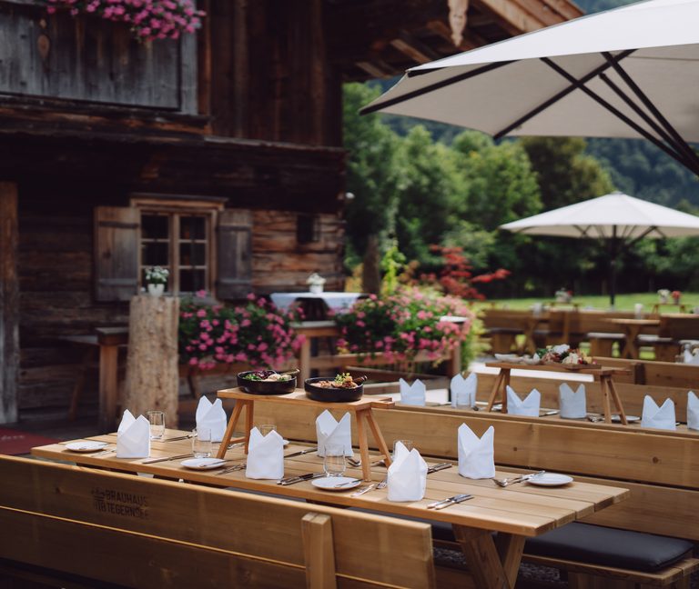 Gedeckte Holztische auf einer Restaurantterrasse vor einem rustikalen Holzhaus mit Blumen und Sonnenschirmen.