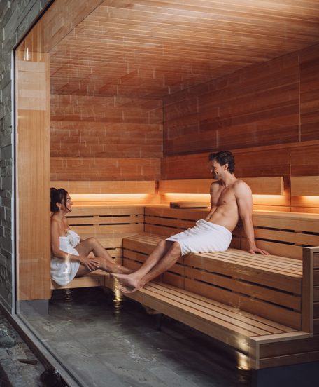 Ein Paar sitzt in einer modernen Sauna mit Holzbank, Glasfront und Steinwänden.