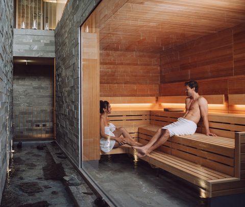 Ein Paar sitzt in einer modernen Sauna mit Holzbank, Glasfront und Steinwänden.