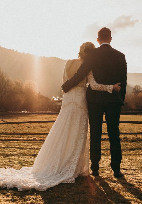 Bride and groom standing in a meadow before mountains, photographed from behind in backlight.