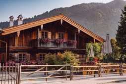 Rustic wooden chalet with balcony, flowers and terrace in front of a forested mountain backdrop.