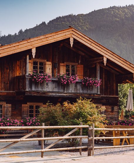 Rustikales Holzchalet mit Balkon, Blumen und Terrasse vor bewaldeter Bergkulisse.