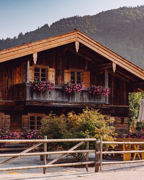 Rustikales Holzchalet mit Balkon, Blumen und Terrasse vor bewaldeter Bergkulisse.