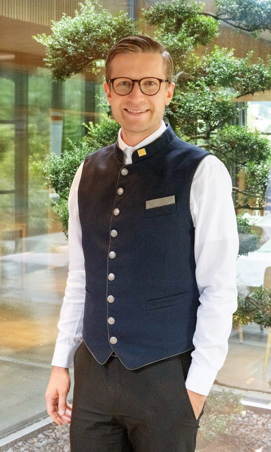 Hotelmitarbeiter in Uniform steht in einem hellen Innenbereich vor einer großen Zierpflanze.