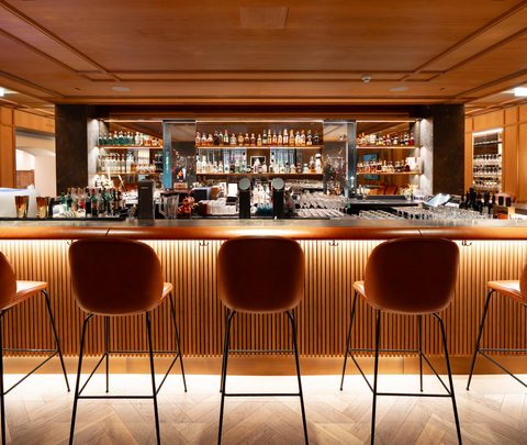 Warm beleuchtete Hotelbar mit Barhockern vor einem Tresen und Regalen mit Flaschen im Hintergrund.