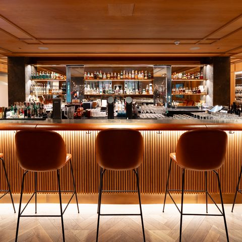 Warm beleuchtete Hotelbar mit Barhockern vor einem Tresen und Regalen mit Flaschen im Hintergrund.