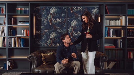 A couple with drinks in a lounge with bookshelves and a leather sofa.