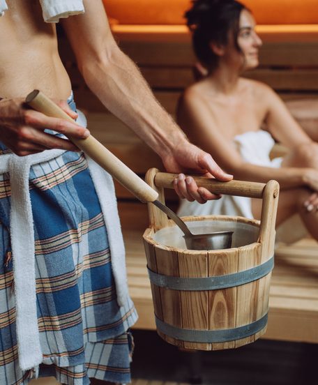 Saunameister mit Holzeimer und Kelle beim Aufguss, Gäste sitzen auf Holzbänken in der Sauna.