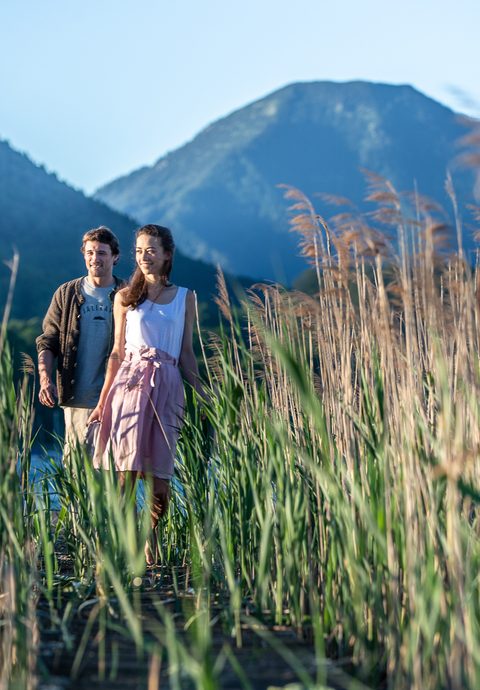 A couple walks through tall reeds with mountains by a body of water.