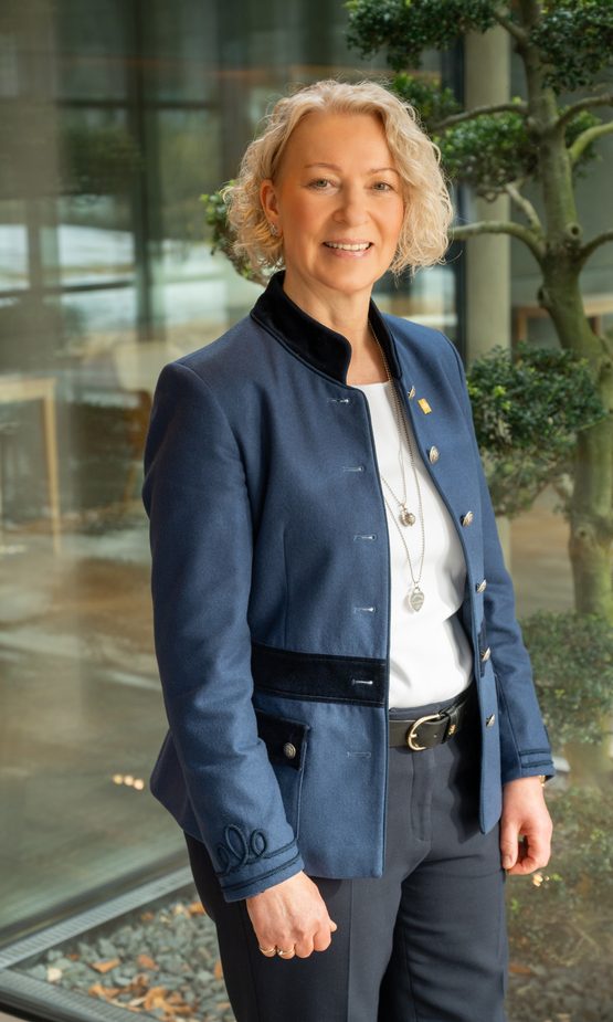 Hotelmitarbeiterin in blauer Jacke steht lächelnd in einer Lobby vor Glasfront und Zierbaum.