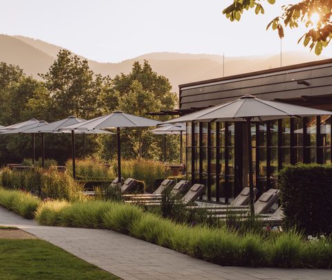 Außenbereich eines Wellnessgebäudes mit Liegen, Sonnenschirmen, Gartenweg und Bergen im Hintergrund.