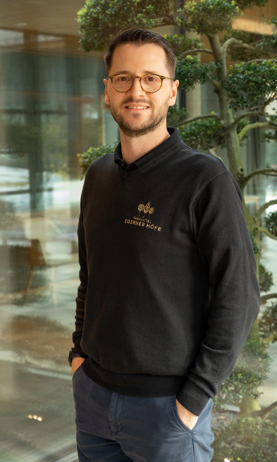 Hotelmitarbeiter mit Brille steht lächelnd in der Lobby vor Glasfront und Zierbaum.