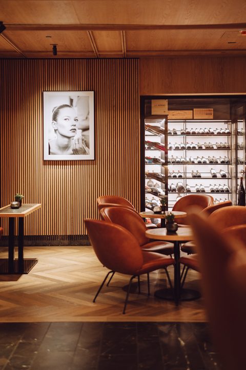 Sitzbereich einer Hotelbar mit Ledersesseln, kleinen Tischen und beleuchtetem Weinschrank an Holzwand.