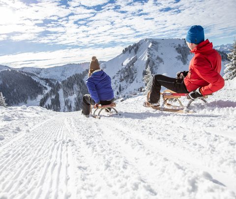 Two people on sleds on a snowy mountain track overlooking snow covered mountains and a cloudy sky.