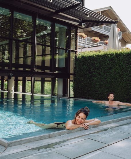 Zwei Gäste entspannen in einem Außenpool neben einem modernen Wellnessgebäude mit Glasfassade.