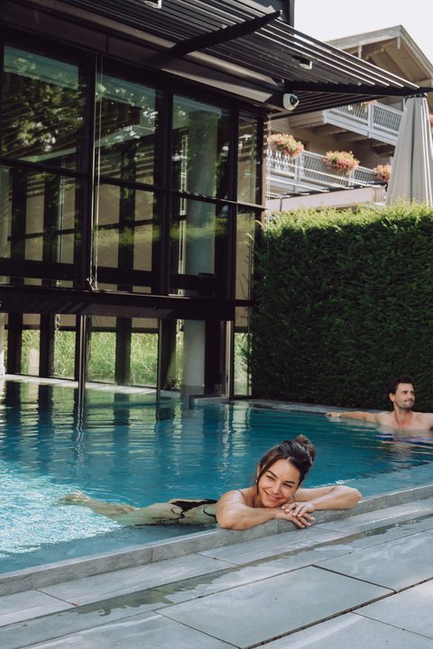 Zwei Gäste entspannen in einem Außenpool neben einem modernen Wellnessgebäude mit Glasfassade.