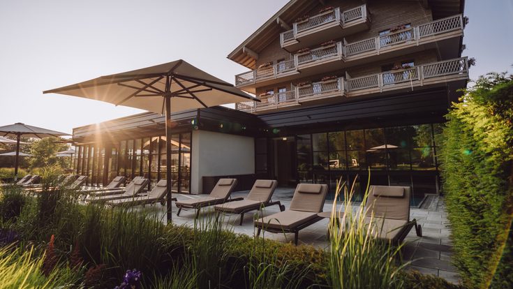 Liegestühle und Sonnenschirme auf der Hotelterrasse vor einem mehrstöckigen Gebäude im Abendlicht.