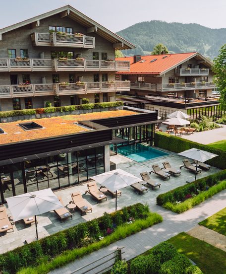 Hotelanlage mit Außenpool, Liegen, Terrasse und Garten vor bewaldeten Bergen.