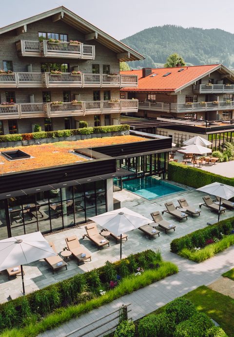 Hotel exterior with outdoor pool, loungers, terrace and garden in front of wooded mountains.