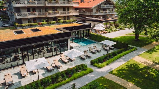 Hotel exterior with outdoor pool, loungers, terrace and garden in front of wooded mountains.