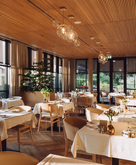 Modernes Restaurant mit gedeckten Tischen, großer Fensterfront, Sitzbank und Holzdecke.