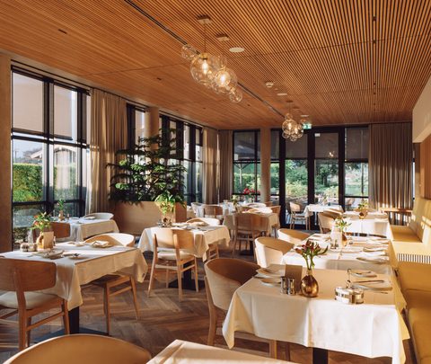 Modernes Restaurant mit gedeckten Tischen, großer Fensterfront, Sitzbank und Holzdecke.