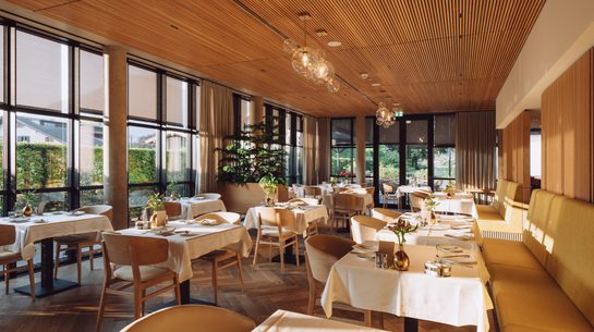 Modern restaurant with set tables, large windows, banquette seating and wooden ceiling.