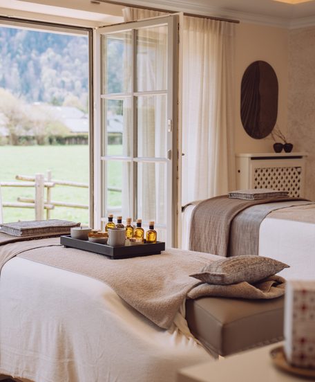 Wellness-Behandlungsraum mit zwei Liegen, Ölen und offenem Fenster mit Blick auf Wiese und Garten.