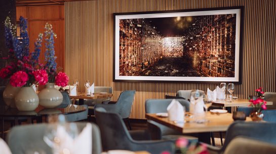 Restaurant interior with set tables, blue armchairs, flower vases, and large wall art on a wood slat wall.
