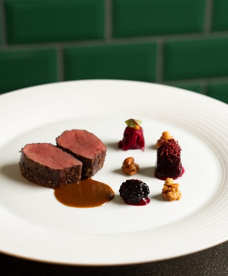 Kunstvoll angerichtetes Gericht mit Fleischmedaillons, Sauce, Brombeere und Rote-Bete-Garnitur auf weißem Teller.