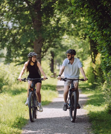 A couple rides bicycles on a path through a green landscape lined with trees.