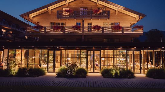 Illuminated chalet-style hotel exterior with balconies, flowers, and a glass ground floor at night.