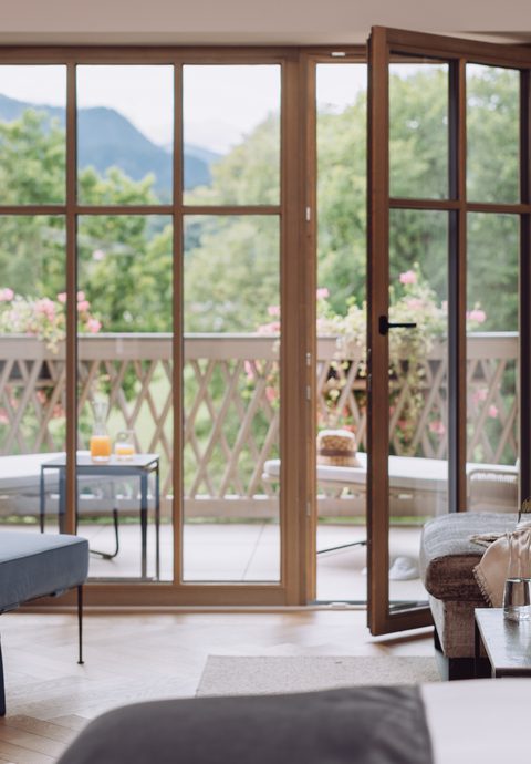 Hotelzimmer mit Sitzbereich, offener Terrassentür und Blick auf Bäume und Berge.