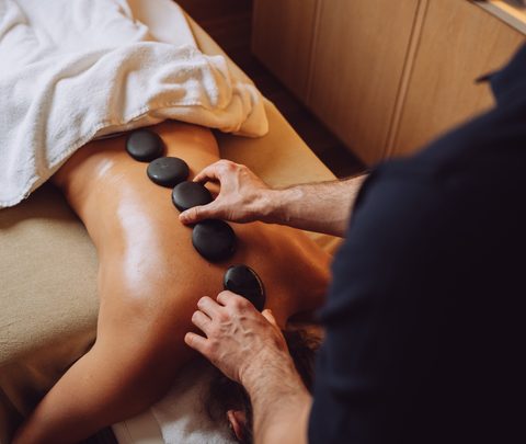 Hot-Stone-Massage mit schwarzen Steinen auf dem Rücken einer Person in einem Wellnessraum.