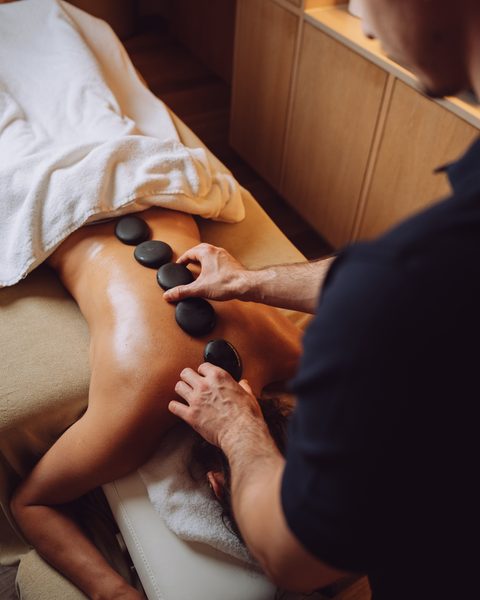 Hot-Stone-Massage mit schwarzen Steinen auf dem Rücken einer Person in einem Wellnessraum.