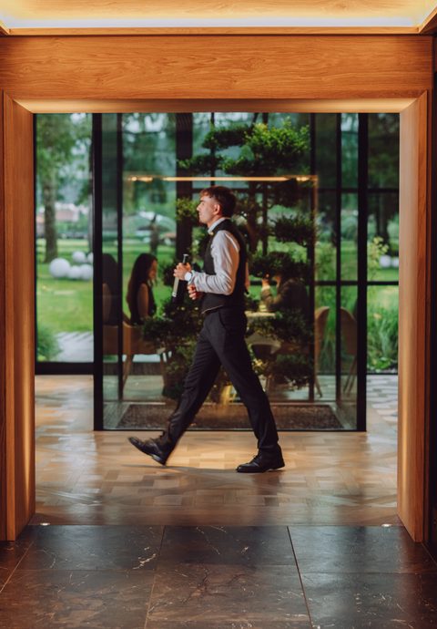 A waiter carries a wine bottle through a corridor lined with glass wine cabinets.