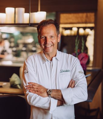 A chef in a white jacket stands with folded arms in front of an open restaurant kitchen.
