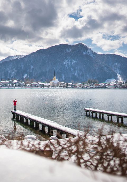 Snow-covered jetties on a lake with a person, village, church and mountains in winter.