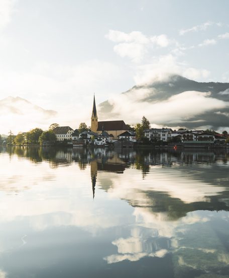 Village with church on a lakeshore reflected in calm water with mountains and clouds behind.