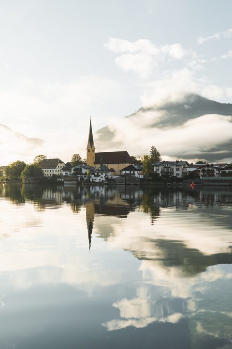 Ort mit Kirche am Seeufer, gespiegelt im ruhigen Wasser vor Bergen und Wolken.