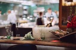 Set restaurant table with glasses, folded napkins and a chilled bottle in front of an open kitchen.
