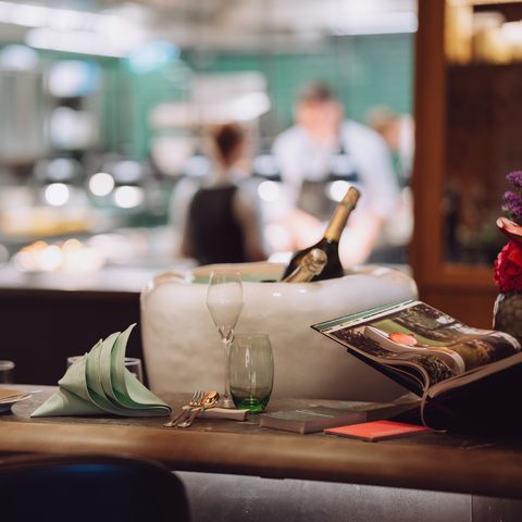 Gedeckter Restauranttisch mit Gläsern, Servietten und gekühlter Flasche vor einer offenen Küche.
