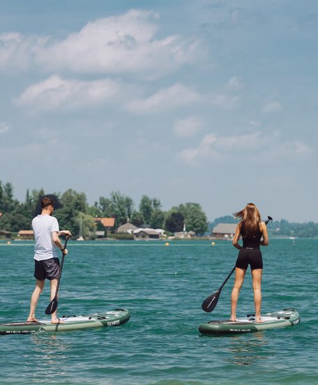 Two people on stand-up paddleboards on a lake, with a shore, trees and houses in the background.