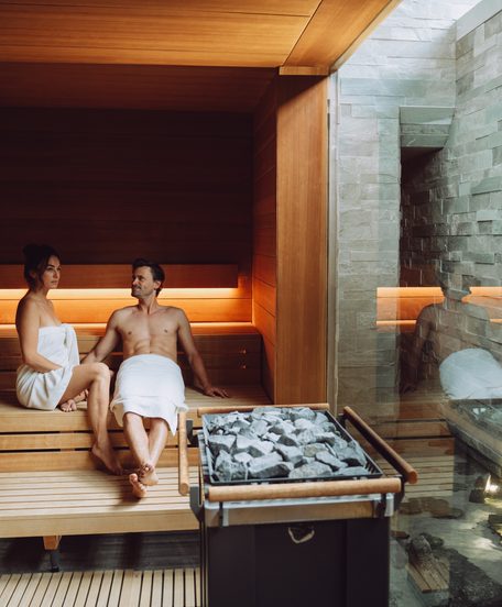 Ein Paar sitzt in einer Sauna mit Holzbank, Saunaofen und Steinwand.