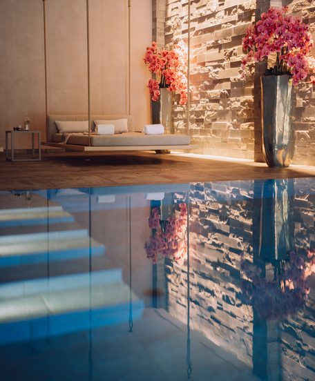 Indoor-Pool im Wellnessbereich mit schwebender Liege, Steinwand und rosa Orchideen.
