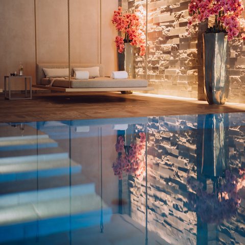 Indoor-Pool im Wellnessbereich mit schwebender Liege, Steinwand und rosa Orchideen.